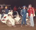1983 Ron Shuman relaxes on the right rear tire of the Pabst Blue Ribbon, Wilke Racer’s winning car as Kevin Olson is interviewed following the November 25 Gardena, CA. Classic. Olson Lead the final 14 laps for his first Turkey Night Trophy. 